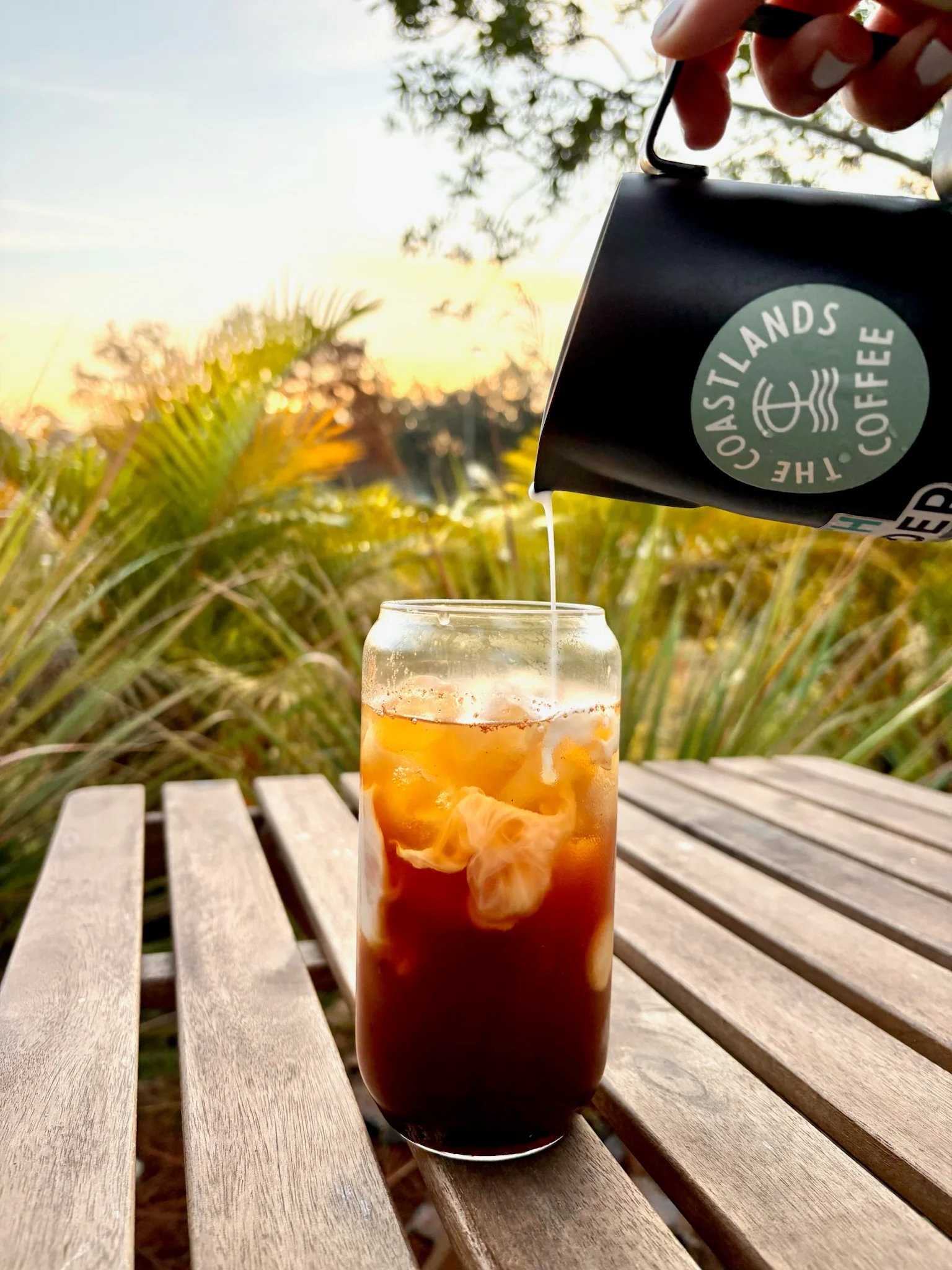 Coffee pouring from a Coastlands carafe into an iced glass outdoors.
