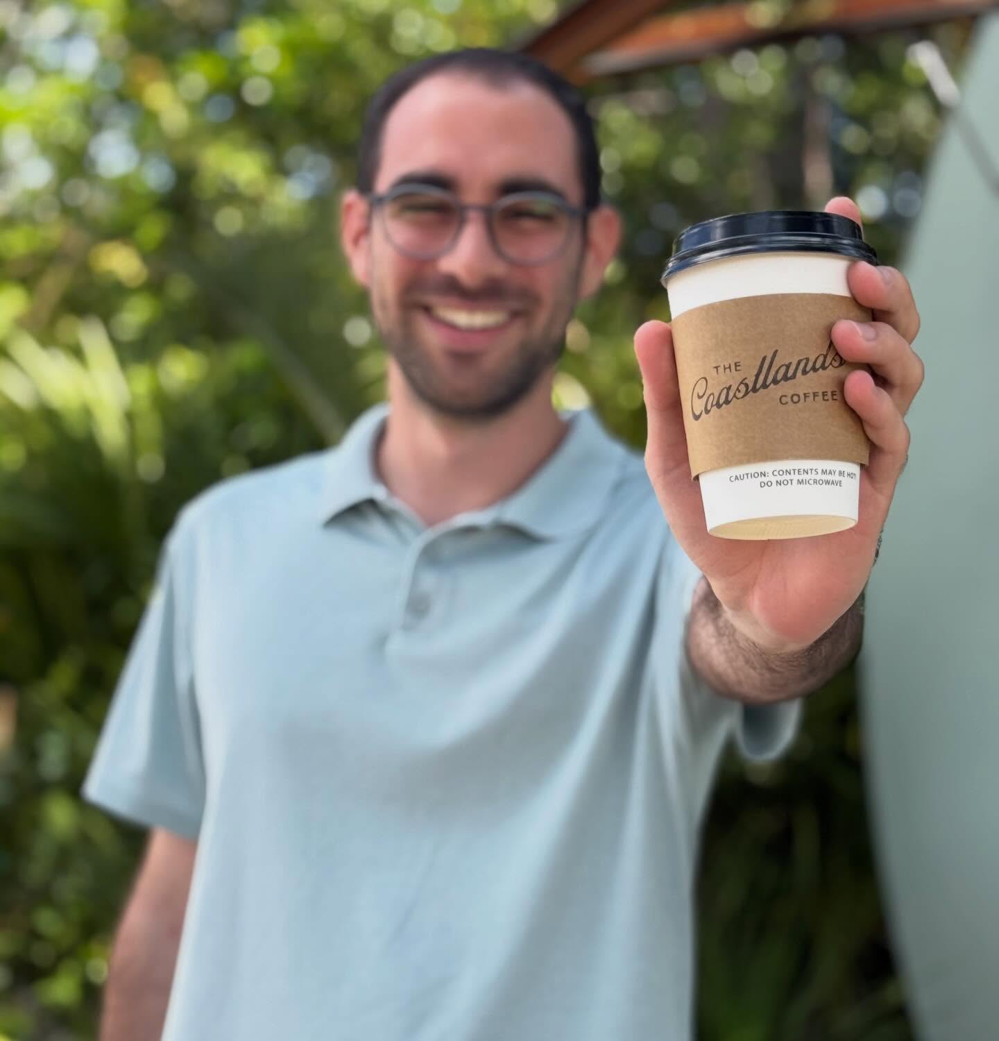 A Coastlands takeaway cup held toward the camera, with a smiling man softly blurred behind it.